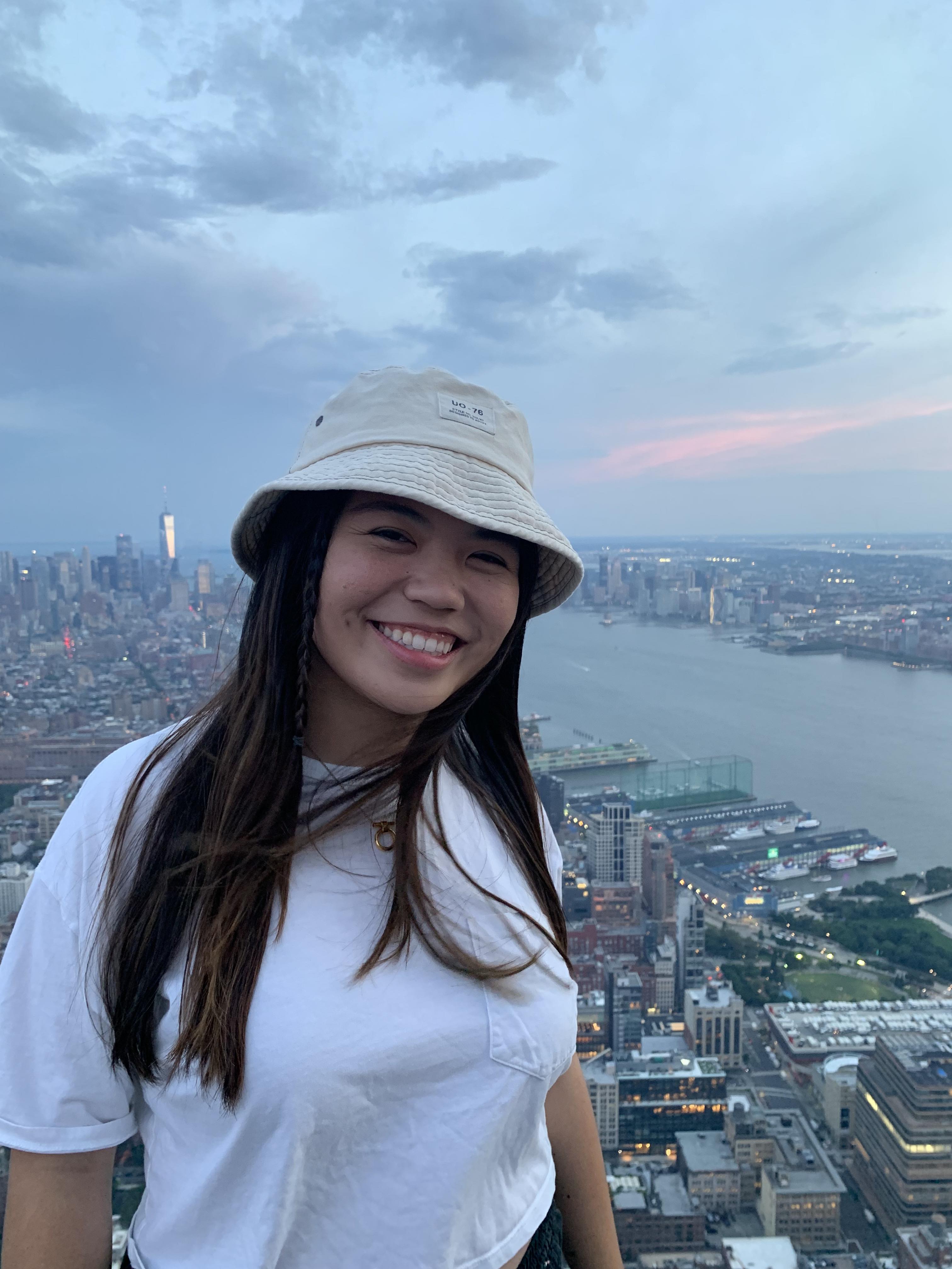 Danielle, a southeast Asian woman with long black-brown hair, wearing a bucket hat with the New York skyline behind her.