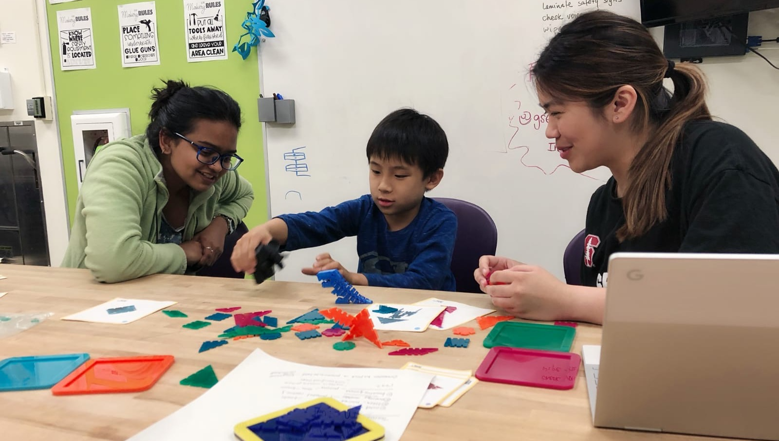 A young Asian boy play-testing with colorful geometric pieces while two women observe.